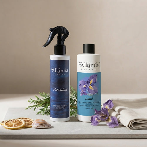 Una fotografia raffinata che presenta due prodotti Alkimia Essence su una lastra di pietra grigia. A sinistra, il flacone spray da 300 ml 'Poseidon' con etichetta blu scuro. A destra, la bottiglia da 200 ml 'Lune' con etichetta azzurro-viola raffigurante un iris. La composizione è minimalista, arricchita da pochi elementi naturali come una piccola conchiglia e un panno di lino, su un fondo neutro.