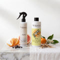 Due bottiglie di Alkimia Essence, 'Eurus' spray per tessuti e 'Aeris' profumatore per bucato, su un piano di marmo con agrumi e ingredienti botanici.