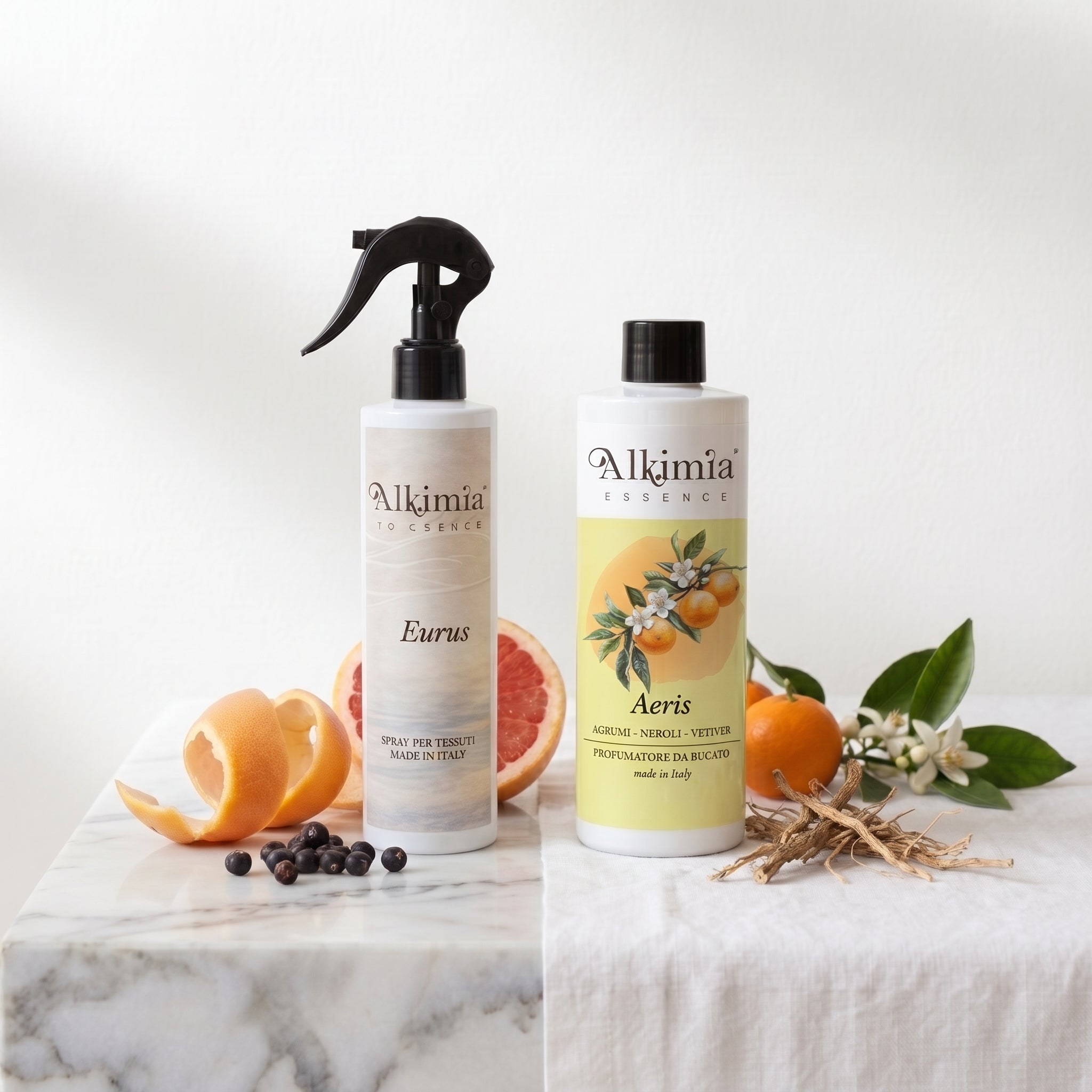 Due bottiglie di Alkimia Essence, 'Eurus' spray per tessuti e 'Aeris' profumatore per bucato, su un piano di marmo con agrumi e ingredienti botanici.